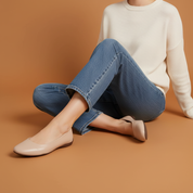 Person wearing a white sweater, blue jeans, and beige flats sitting on a brown background