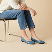Person wearing blue flats and jeans sitting on a chair against a beige background